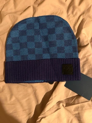 Blå Louis  mössa med rutmönster - Snygg blå mössa från Louis med klassiskt rutmönster i två nyanser av blått. Mössan har en ribbstickad uppvikt kant och en svart patch med Louis -logga framtill. Perfekt accessoar för dig som vill sticka ut med stil.