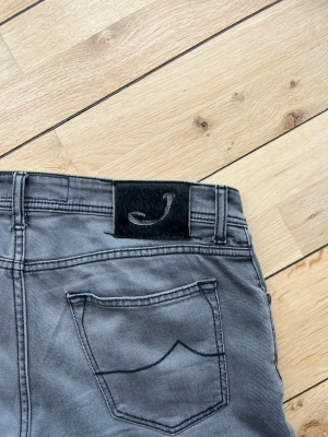 Grå jeans från Jacob Cohën - Snygga grå jeans från Jacob Cohën med raka ben och klassiska bakfickor. Jeansen har en svart patch med ett J på linningen och är gjorda i mjukt denimtyg. Perfekta för en avslappnad men stilren look.