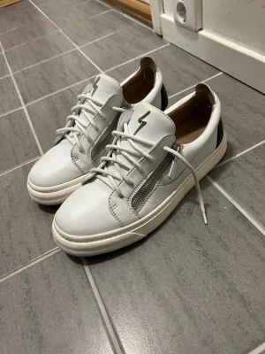 Gisueppe Zanotti - Snygga vita sneakers med svart detalj på hälen, blixtsymbol på plösen och dragkedja på sidan. Skorna har vita snören och en tjock vit sula. Tillverkade i slätt läderliknande material och har en modern, clean look.