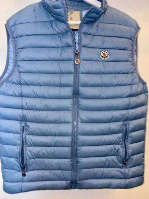 Ljusblå dunväst - Snygg ljusblå dunväst med quiltad design och två dragkedjefickor framtill. Västen har hög krage, hel dragkedja och logga på bröstet. Perfekt för lager-på-lager och stilren look.