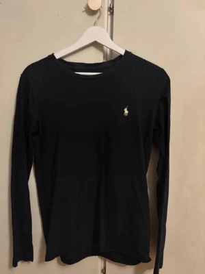 Svart långärmad topp Polo Ralph Lauren - Klassisk svart långärmad topp från Polo Ralph Lauren med rund halsringning och den ikoniska loggan broderad på bröstet. Tillverkad i mjuk bomull som känns skön mot huden. Perfekt basplagg till garderoben.