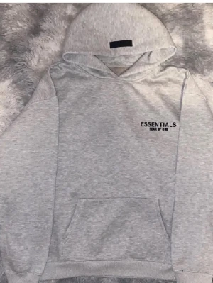  Ljus Grå Essentials Fear of God hoodie - Snygg ljusgrå hoodie från Essentials Fear of God med svart logga på bröstet och stor text på ryggen. Klassisk känguruficka framtill och stor huva. Tillverkad i mjukt bomullsmaterial som är perfekt för en chill och avslappnad stil.