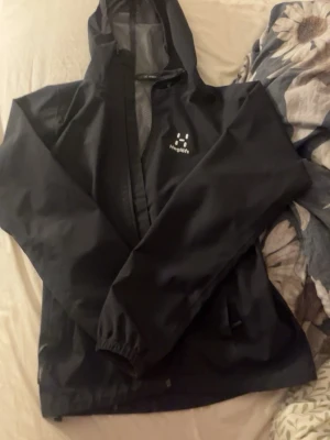 Svart regnjacka från Haglöfs - Snygg svart regnjacka från Haglöfs med luva och dragkedja framtill.  En jacka för tjejer, inget fel med den! Xs-S