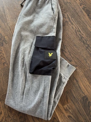 Grå mjukisbyxor Lyle & Scott - Snygga grå mjukisbyxor från Lyle & Scott med svart cargoficka på benet och gul logga. Byxorna har ribbade muddar vid benslut och dragsko i midjan. Perfekta för chill dagar och streetwear-stil.