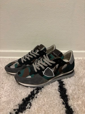 Philippe Model TRPX camo - Snygga sneakers från Philippe Model med camo-mönster i grönt, svart och brunt. Skorna har grå mockadetaljer, vita och grå partier samt vita snören. Yttermaterialet är en mix av mocka och textil, med svart sula och logga på sidan.