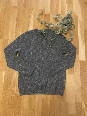 Gråmelerad stickad tröja Lyle & Scott - Snygg gråmelerad stickad tröja från Lyle & Scott med rund halsringning och klassisk gul logga på bröstet. Tröjan har långa ärmar och ribbade muddar vid ärmslut och nederkant. Perfekt för lager-på-lager och stilren vardagslook.