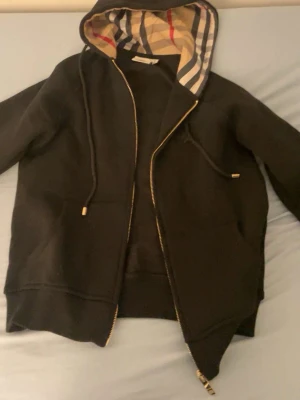 Svart hoodie med Burberry-rutor - Snygg svart hoodie från Burberry med dragkedja och klassiskt rutigt foder i huvan. Hoodien har dragsko vid huvan och ribbade muddar. Perfekt för dig som gillar stilrena plagg med en lyxig touch. Materialet känns mjukt och bekvämt.