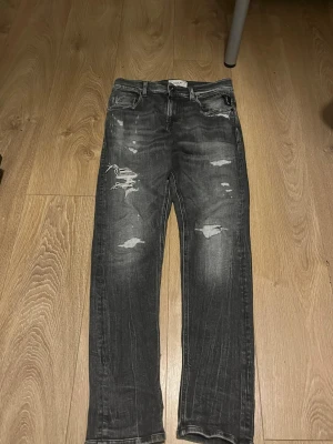Svarta slitna jeans med raka ben - Säljer ett par svarta jeans med slitningar och distressed detaljer på både fram- och baksida. Jeansen har raka ben, klassisk femficksmodell och cool tvättad look. Perfekta för dig som gillar en avslappnad och trendig stil.