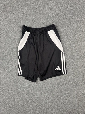 Svarta Adidas träningsshorts - Snygga svarta träningsshorts från Adidas med vita detaljer och klassiska ränder på sidorna. Shortsen har elastisk midja med snörning och är tillverkade i ett lätt och snabbtorkande syntetmaterial. Perfekta för sport eller gym!