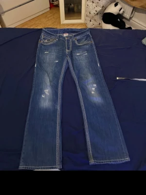 Blå True Religion bootcut jeans - Säljer ett par blå bootcut jeans från True Religion med vita kontrastsömmar och slitna detaljer framtill. Klassisk femficksmodell och tillverkade i USA. Perfekta för dig som gillar en avslappnad men ändå snygg stil.