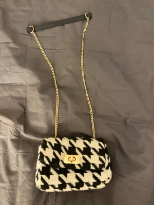 Houndstooth handväska med guldkedja - Snygg handväska i svartvitt houndstooth-mönster med guldfärgat vridlås och kedjerem med svart axelband. Väskan har en mjuk, fluffig textur och klassisk rektangulär form. Perfekt accessoar för att lyfta din outfit.