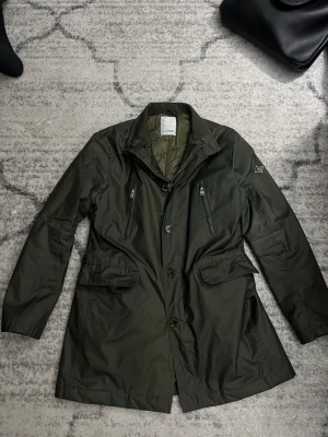 Mörkgrön rock från J.Lindeberg - Stilren mörkgrön rock från J.Lindeberg med två bröstfickor med dragkedja och två stora fickor med lock. Rocken har klassisk krage, diskret logotyp på ärmen och är tillverkad i ett glansigt, vattenavvisande material. Perfekt för en trendig och funktionell look.