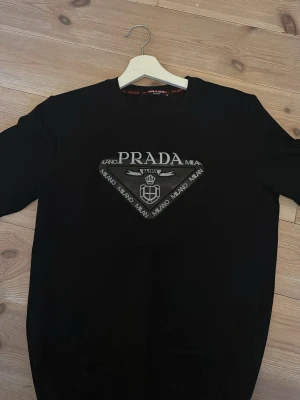 Svart tröja från Prada Milano - Snygg svart tröja från Prada Milano med stort logotryck på bröstet. Rund halsringning och klassisk passform. Perfekt för dig som gillar exklusiva märken och stilren design.