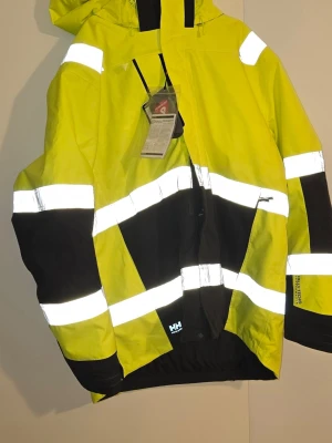 Gul Helly Hansen vinterjacka 4XL  - Snygg gul vinterjacka från Helly Hansen med svarta detaljer och breda reflexband runt ärmar, bröst och midja. Jackan har huva, dragkedja och praktiska fickor. Perfekt för dig som vill synas och hålla dig torr i alla väder.