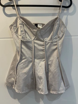 Champagnefärgad korsettopp med smala band - Snygg korsettopp i champagnefärgat satinliknande material med smala axelband och markerade sömmar. Toppen har utsvängd nederdel och smock i ryggen för skön passform. Perfekt för att lyfta vilken outfit som helst.