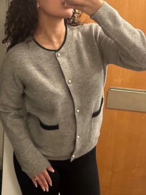 Grå kofta med svarta detaljer - Säljer en grå kofta med svarta kanter vid fickorna och halsringningen. Koftan har pärlknappar framtill och två fickor. Den är stickad och har en klassisk, rak passform med långa ärmar. Perfekt för lager-på-lager och enkel att matcha.