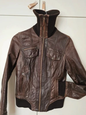 Brun vintage skinnjacka med muddar - Snygg brun skinnjacka i läder med ribbade muddar och hög stickad krage. Jackan har dragkedja framtill, två bröstfickor med lock och knappar samt två snedställda fickor. Perfekt för dig som vill ha en cool och tidlös look.