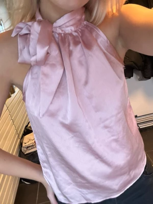 Rosa halterneck topp med rosett - Snygg ljusrosa halterneck topp i glansigt satinliknande material. Toppen är ärmlös och har en stor rosett vid halsen som ger en extra touch. Perfekt för dig som vill sticka ut lite extra.
