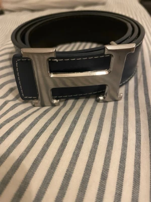 Svart läderbälte med H-spänne - Stilrent svart läderbälte från H med ikoniskt H-format metallspänne i silverfärg. Bältet har vita kontrastsömmar längs kanterna och passar perfekt för att lyfta din outfit med en lyxig touch.