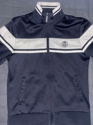 Svart vindjacka från Sergio Tacchini - Sportig svart vindjacka från Sergio Tacchini med vita ränder och perforerade detaljer över bröst och ärmar. Jackan har hög krage, dragkedja framtill och två sidofickor. Perfekt för en streetwear-look.