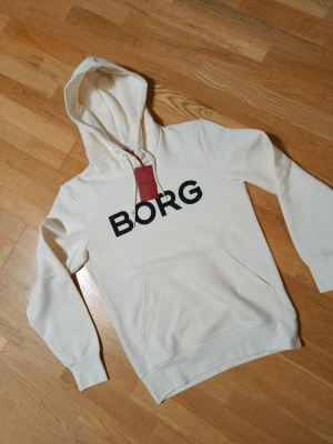 Vit hoodie från BORG med tryck - Snygg vit hoodie från BORG med stor svart logga på bröstet. Klassisk modell med huva och dragsnören samt en stor magficka framtill. Tillverkad i mjukt material som känns skönt mot huden. Perfekt för en avslappnad och trendig stil.