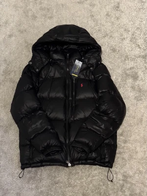 Svart dunjacka från Polo Ralph Lauren - Snygg svart dunjacka från Polo Ralph Lauren med huva och dragkedja. Jackan har en klassisk puffer-design, justerbara snören i nederkant och liten röd logga på bröstet. Perfekt för kalla dagar och riktigt skön att bära.