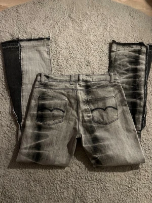 grå jeans med unik tvätt - Säljer ett par grå jeans från Replay med coolt svart tvättat mönster och råa kanter längst ner på benen. Klassisk femficksmodell med svarta detaljer och logga på insidan av linningen. Perfekta för dig som vill sticka ut med din stil.