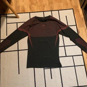 Svart långärmad träningströja Gymshark - Svart långärmad träningströja från Gymshark med röda detaljer och mönster över axlar och armar. Tröjan har en tight passform och är tillverkad i ett stretchigt funktionsmaterial som andas. Perfekt för gym eller löpning.