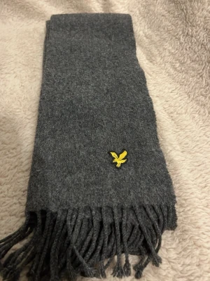 Mörkgrå halsduk från Lyle & Scott - Mysig mörkgrå halsduk från Lyle & Scott med klassisk gul broderad örnlogga. Halsduken är i mjukt material och har fransar i ändarna för en stilren look. Perfekt accessoar för att hålla värmen och lyfta din outfit. Priset kan diskuteras vid snabb affär🫱🏻‍🫲🏽