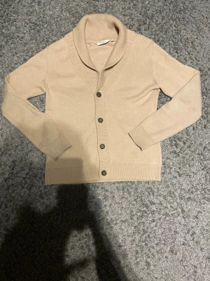 Cardigan  - Stilren beige cardigan med sjalkrage och fem knappar framtill. Cardigan  är ribbstickad i mjukt material och har långa ärmar samt ribbade muddar vid ärmslut och nederkant. Perfekt för lager-på-lager och enkel att matcha.