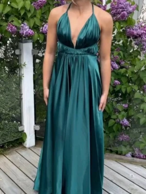 Grön långklänning i satin - Elegant långklänning i smaragdgrön satin med djup v-ringning och tunna axelband. Öppen i ryggen, varierar i storlek då man kan knyter banden själv🥰🥰