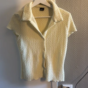 Ljusgul kortärmad skjorta Gina Tricot - Kortärmad skjorta från Gina Tricot i ljusgul färg med strukturerad, stickad känsla. Skjortan har klassisk krage, knappar framtill och lätt vågig kant nertill. Perfekt för dig som gillar en retrovibe och vill ha något unikt i garderoben. Ser större/bredare ut på bild 