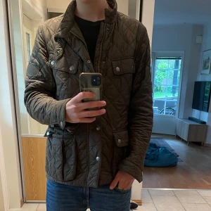 Quiltad höstjacka från Barbour M - Säljer en grön quiltad jacka från Barbour i storlek M. Jackan har snyggt rutmönster, flera fickor med tryckknappar och dragkedja framtill. Insidan har ett coolt tryck med motorcykelmotiv. Perfekt för dig som gillar klassisk stil med edge.