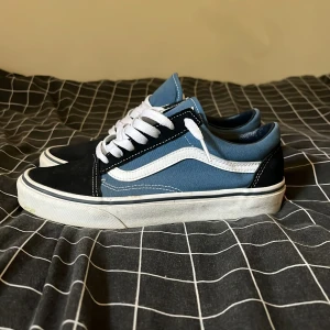 Vans Old Skool blå och svarta sneakers - Säljer ett par klassiska Vans Old Skool sneakers i blått och svart med vit sula och vit sidestripe. Skorna har snörning, rund tå och är tillverkade i canvas och mocka. Perfekta för dig som gillar streetstyle och vill ha en tidlös look.