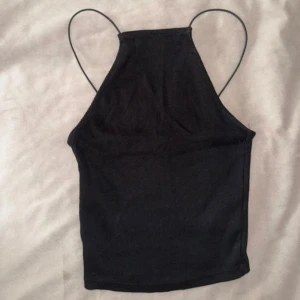 Svart ribbad linnetopp med tunna band - En svart ribbad linnetopp med högt skuren hals och tunna axelband. Toppen har en rak passform och ärmlös design, perfekt för fest, varma dagar eller lager-på-lager. Enkel och stilren look som funkar till många outfits.