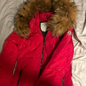 Röd Moncler dunjacka med päls - Säljer en röd Moncler dunjacka med stor fluffig päls runt kragen. Jackan har dragkedja framtill, svarta detaljer och klassisk Moncler-logga på ärmen. Perfekt för kalla dagar och riktigt snygg färg som sticker ut.