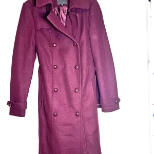  kappa från Amisu med knappar - Snygg kappa från Amisu med dubbelknäppning och dekorativa knappar på axlar och ärmslut. Lång modell med klassisk krage och fodrad insida. Perfekt för dig som vill ha en stilren och tidlös look under höst och vinter.