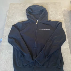 Svart hoodie från GANT med dragkedja - Svart hoodie från GANT med broderad logga och texten 'GANT 1949' på bröstet. Tröjan har huva, hel dragkedja framtill och två fickor. Tillverkad i mjuk bomullsmix, perfekt för chill dagar eller när du vill ha en clean look.