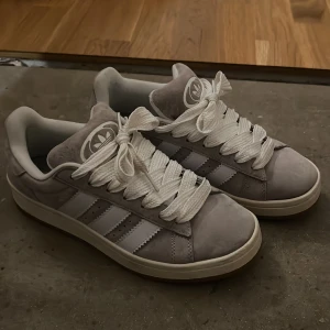 Adidas campus i grå mocka, strl 39⅓ - Snygga Adidas campus i ljusgrå mocka med klassiska vita ränder på sidorna och breda vita skosnören. Skorna har rund tå, platt sula och Adidas logga på plösen. Använda ett fåtal gånger. Som nya!