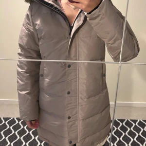 Beige dunjacka från Everest med huva - Säljer en beige dunjacka från Everest i storlek 38. Jackan har huva med fuskpäls, dragkedja och knappar framtill samt två stora fickor. Perfekt för kalla dagar och riktigt skön att bära. Klassisk lång modell som håller värmen.