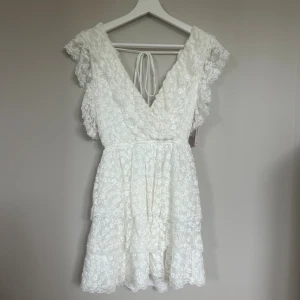 Vit spetsklänning från Nelly XXS - Vit spetsklänning från Nelly med omlott och v-ringad fram och bak. Klänningen har volangdetaljer på axlarna, knyt i ryggen och är kort i modellen. 