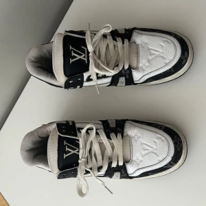 Louis Vuitton svartvita sneakers - Säljer ett par Louis Vuitton sneakers i svart och vitt med klassisk LV-logga på tungan och hälen. Skorna har snörning, rund tå och platt sula. Ovandelen är i skinn med monogramdetaljer i textil och kontrasterande paneler. Snygg och lyxig design som sticker ut.