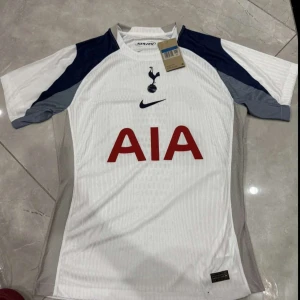 Tottenham Hotspur fotbollströja Nike - Snygg Tottenham Hotspur matchtröja med Kudus 20 på ryggen. Tröjan är vit med mörkblå och grå detaljer på ärmarna, röd AIA-logga på bröstet och klubbens emblem. Tillverkad i lätt och ventilerande material, perfekt för fotboll eller häng med kompisar. 10/10. Helt ny. Aldrig använd.