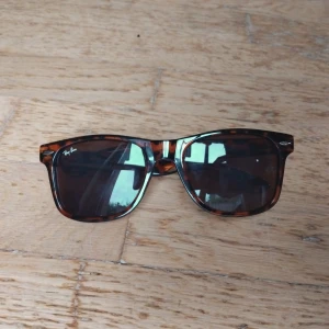 Ray-Ban Wayfarer leopardmönstrade solglasögon - Snygga Ray-Ban Wayfarer solglasögon med leopardmönstrad båge i brunt och orange. Glasen är mörka och rektangulära, och bågen är tillverkad i plast. Ikonisk Ray-Ban-logga på linsen och insidan av skalmen. Perfekt för dig som vill sticka ut med klassisk stil.
