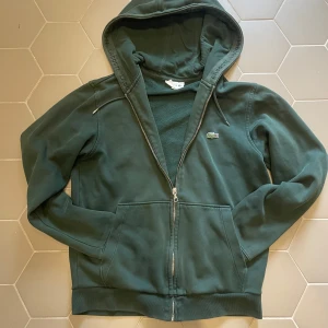 Grön hoodie från Lacoste med dragkedja - Mörkgrön hoodie från Lacoste med dragkedja och huva. Klassisk krokodillogga på bröstet och diskret Lacoste-tryck på insidan av huvan. Mjuk bomullskänsla och två fickor framtill. Perfekt för chill dagar eller när du vill ha en clean look.