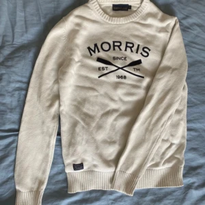 Vit stickad tröja från Morris - Vit stickad tröja från Morris R. Club med svart tryck framtill och ribbade muddar. Klassisk rund halsringning och långärmad modell. Snygg logga med korsade åror och texten 'Morris Since 1968' på bröstet. Perfekt för dig som gillar stilrena och sportiga plagg.