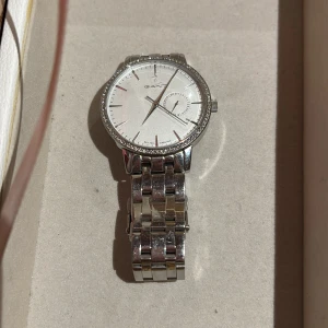 Silverfärgad klocka från GANT - Stilren klocka från GANT med rund urtavla i silver och diskreta indexmarkeringar. Boetten har en glittrig kant och armbandet är i polerat metall med länkdesign. Urtavlan har en extra liten visare för datum. Perfekt för dig som gillar klassisk och clean stil.