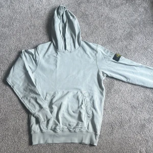 Ljusgrå hoodie från Stone Island - Snygg ljusgrå hoodie från Stone Island med klassisk patch på ärmen. Tröjan har huva, känguruficka och ribbade muddar. Perfekt för dig som gillar streetwear och vill ha en stilren look.