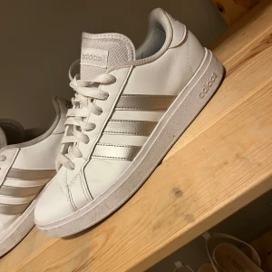 Vita Adidas sneakers med silvriga ränder - Snygga vita Adidas sneakers med klassiska silvriga ränder på sidorna och vit sula. Skorna har snörning och är tillverkade i syntetmaterial med en stilren och sportig look. Perfekta för dig som gillar en clean och tidlös stil. Använda högst 3ggr 