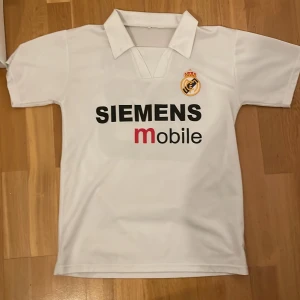 Real Madrid Beckham 23 fotbollströja - Klassisk vit Real Madrid fotbollströja med svart Siemens Mobile-tryck på bröstet och broderat klubbmärke. På ryggen står det Beckham och nummer 23 i svart. Tröjan har krage och är tillverkad i lätt polyestermaterial, perfekt för fotbollsälskare.